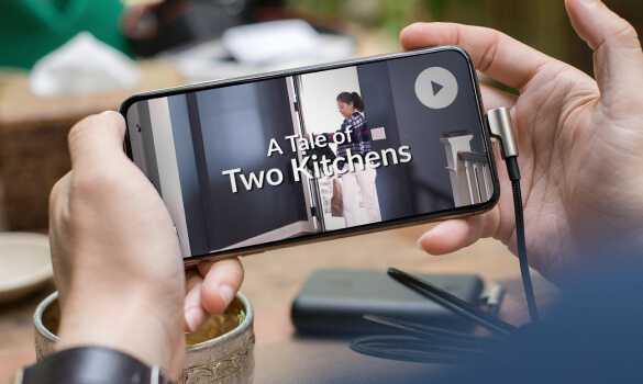 A person holding a smartphone which has a screen which reads, "A Tale of Two Kitchens."