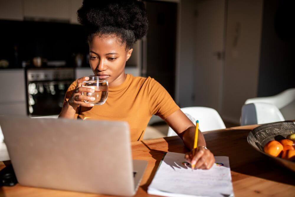 A woman raising a glass of water to her mouth while looking at a laptop.