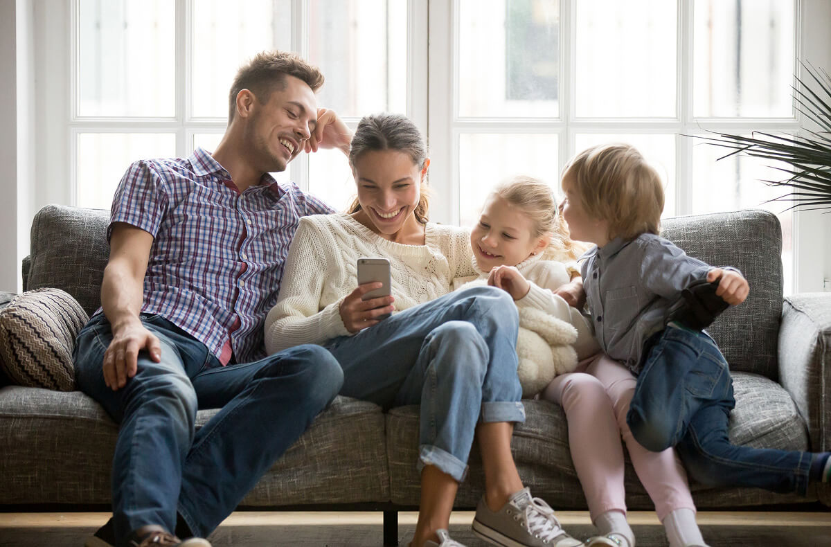 A family of four sitting on a couch and smiling while looking at a cellphone.