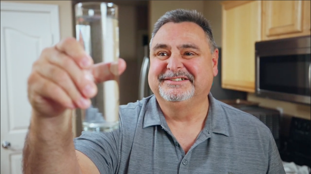 Man holding a glass of water in front of his smiling face.